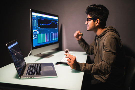 Young indian man pointed on coin looking on display with bitcoin cryptocurrency market in dark office