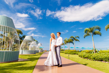 Beautiful blonde bride in white wedding dress and the groom on the roof of the hotel. Tropical sea and palm trees in the background. Summer vacation concept.