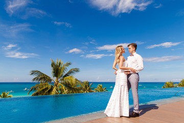 Beautiful blonde bride in white wedding dress and the groom dance in a hotel near the infinity pool. Tropical sea, sky and palm trees in the background. Summer vacation concept.