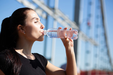 Lovely brunette fit girl drinking water during jogging at the bridge in sunny morning. Space for text