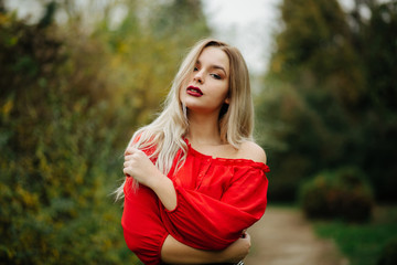 Gorgeous blonde woman in red blouse posing with naked shoulder at the thuja trees
