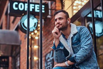 A pensive stylish man wearing a denim jacket with wireless headphones holding takeaway coffee...