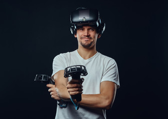 Portrait of a man with bristle dressed in white shirt wearing virtual reality glasses and holds joysticks, looking at camera.