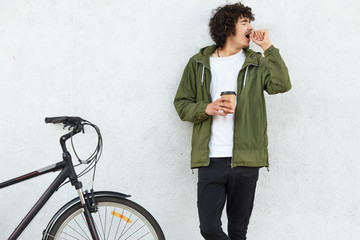 Image of handsome male yawns as wants to sleep, keeps hands near mouth, drinks strong hot coffee to feel refreshed, being tired after riding sports bicycle, isolated over white concrete wall. © sementsova321