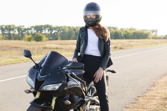 Street style concept. Active professional rider wears helmet, black leather jacket, poses on motorrbike at road, enjoys high speed, ready to cover long distance and have trip. Horizontal shot