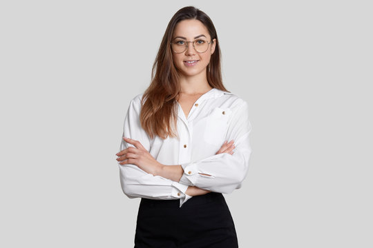 Cheerful Self Confident Businesswoman Wears White Blouse And Black Skirt, Keeps Arms Folded, Wears Round Spectacles, Poses Against White Background, Listens Interlocutor, Dressed In Formal Clothes