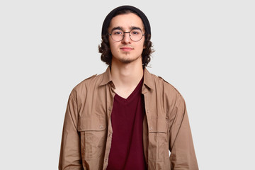 Photo of curly male teenager wears round optical spectacles, black hat, casual shirt, looks...