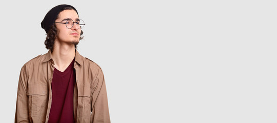 Horizontal shot of pensive young hipster looks pensively aside, wears round spectacles, casual clothes, isolated over white background with free space for your advertising content or promotion