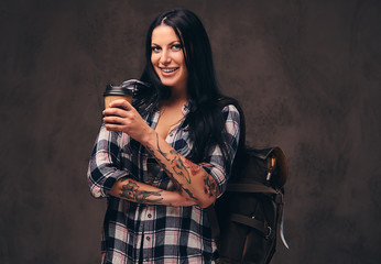 Smiling tattooed girl backpack holding a takeaway coffee in a studio.