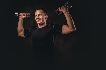 Naklejka na ściany i meble A young bodybuilder wearing sportswear doing exercise with dumbbells on dark background.