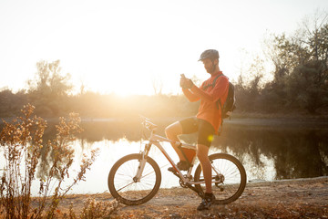 Fototapeta premium Smiling young man sitting on a mountain bike by the lake, resting and using a smart phone. Healthy and active lifestyle concept