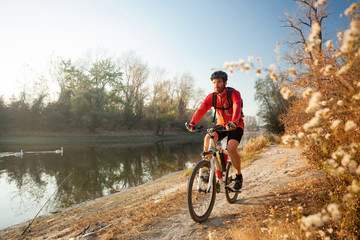 Determined young man riding mountain bike on a footpath along the lake or river on a clear autumn day. Healthy and active lifestyle concept