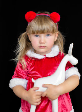 Little beautiful girl in Christmas Santa costume, with fur balls on her head, holding a deer in ruach. The concept of celebrating Christmas, New Year and gifts.
