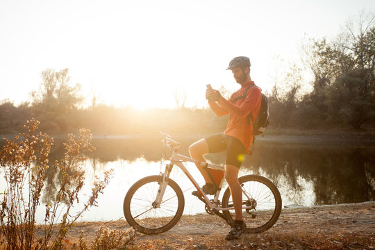 Smiling young man sitting on a mountain bike by the lake, resting and using a smart phone. Healthy and active lifestyle concept