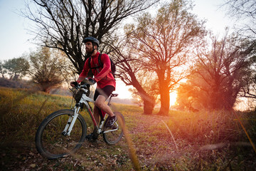 Fototapeta na wymiar Determined young man exercising, riding mountain bike along a footpath through forest. Sunset on a beautiful autumn day. Healthy and active lifestyle concept