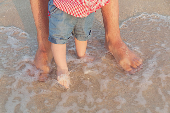 Man And Baby Feet Standing In Shallow Water Waiting For The Wave. Bare Feet Father And His Little Daughter Or Son Staying In The Sand Near The Sea. Concept Of Travel And Holidays. Toned. Soft Focus.