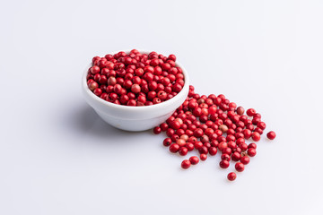 White bowl of Pink Pepper with wooden spoon, Brazil Pepper, Rose pepper, Schinus terebinthifolius seeds over white background, angle view