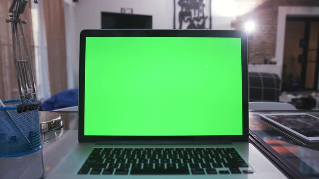 Chrome key green screen. Close-up of screen of silver laptop with black keyboard. Living room with gadgets on table. Indoors. Lifestyle. Evening. Chrome key green screen