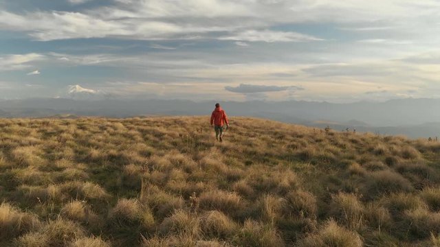 An aerial view of a bearded hipster male with a remote from a drone walks along the edge of a high plateau near a cliff at sunset. The guy walks along the cliff
