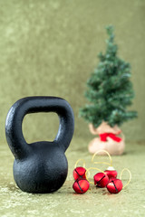 Naklejka na ściany i meble Black kettlebell on a green velvet background with red jingle bells on gold thread, holiday fitness, Christmas tree in background
