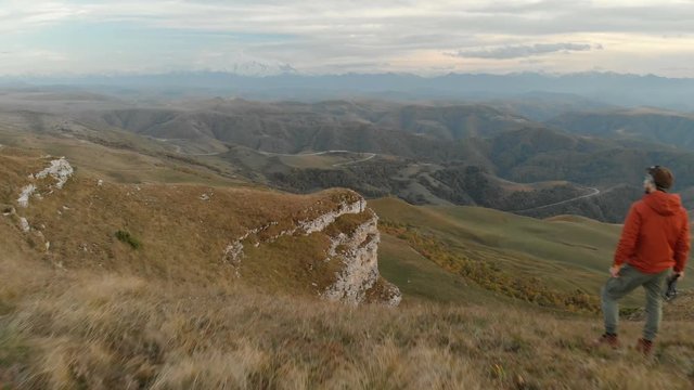 An aerial view of a bearded hipster male with a remote from a drone walks along the edge of a high plateau near a cliff at sunset. The guy is on the cliff