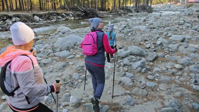 Woman do Nordic walking in nature near mountain river. Girls and children use trekking sticks and nordic poles, backpacks. Family travels sports. Kid is learning from mother and grandmother the proper