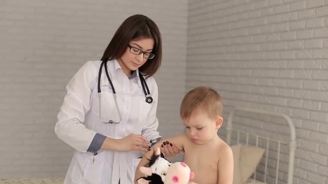 Doctor giving a child an intramuscular injection in arm. Young woman pediatrician performs a vaccination of a little boy. The boy is holding his favorite toy. Vaccination concept.
