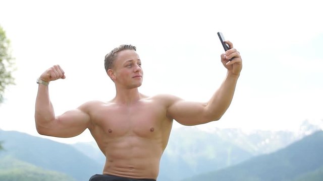 Young athlete half naked caucasian blonde man making photos of his well-trained strong body over beautiful mountains landscape to promote and advertise sports and healthy lifestyle.