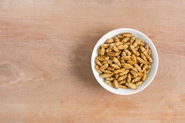 top view of roasted silkworm pupae with salt and soy sauce in a ceramic dish on wooden table, copy space. alternative high nutrition food concept.