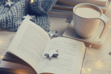 Winter books. Winter cozy reading.  book,  gray knitted scarf,  cup of coffee, shining garland and silvery decorative stars on a light background.
