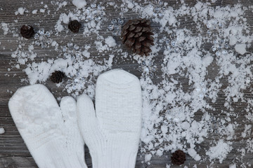 white mittens on the background of snow-covered wood surfaces with pine cones and snowflakes