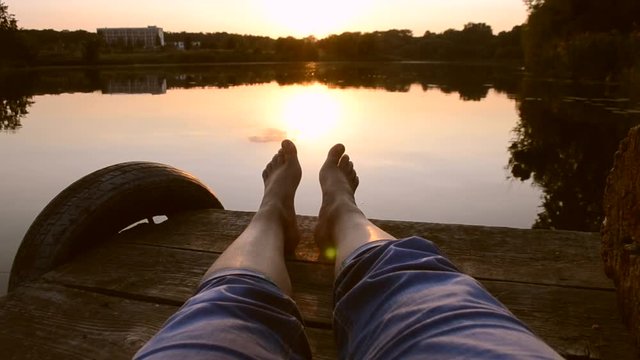 Feet legs lie on pier near lake pond at sunset dawn sunrise close-up. Smooth water surface on which the sun is reflected, windless weather. Black strip of the horizon. POV, point of view. Wide angle.
