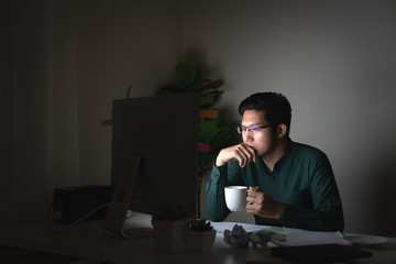 Attractive young asian man drinking coffee sitting on desk table looking at laptop computer in dark late night working feeling serious thinking and determinated at home office in work hard concept.