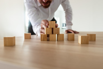Businessman leaning in to carefully assemble pyramid shape with blank wooden blocks