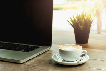 Coffee cup with laptop computer on wooden table and morning light.