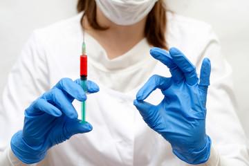 a woman in a white robe, mask and blue gloves, holding a syringe with red liquid