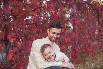 Dad and little son walk in the autumn park. Bright, warm autumn. Red leaves. Cozy. Portrait of dad and son in knitted sweaters.