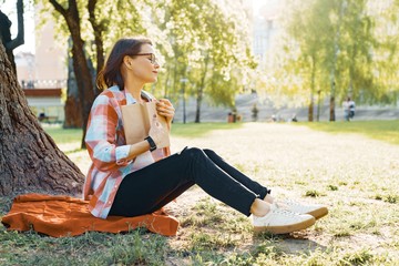 Beautiful mature woman with glasses reading book sitting on the grass near the tree in the park at sunset.