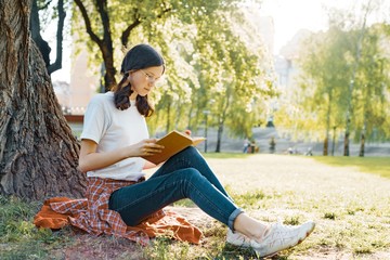 Girl student in glasses reading a book in the park sitting under a tree on the grass