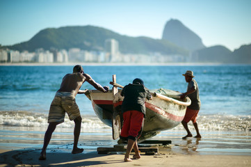 Fishermen pushing a traditional wooden fishing boat into the sea at Copacabana Beach with the dramatic mountain skyline of Rio de Janeiro, Brazil