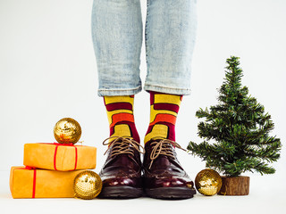 Men's legs, funny socks, stylish shoes, gifts with a red ribbon and a Christmas tree. White background, isolated, close-up. Preparing for the holidays. Сoncept of fashion and elegance