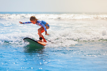 Happy baby girl - young surfer ride on surfboard with fun on sea waves. Active family lifestyle, kids outdoor water sport lessons and swimming activity in surf camp. Beach summer vacation with child.