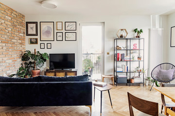 Stylish scandinavian open space with dining and living room with design sofa, bookstand and armchair. Bright and sunny room with plants, poster wall and brown wooden parquet.	