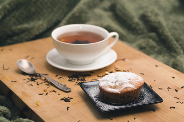 Homemade cupcake with powdered sugar on a black plate and a white cup of tea with natural additives on a wooden tray on a background of green textiles close up. The concept of a cozy breakfast in bed