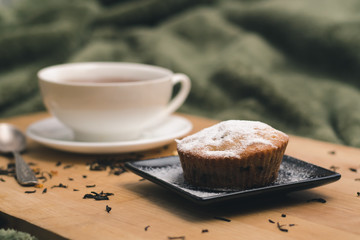 Homemade cupcake with powdered sugar on a black plate and a white cup of tea with natural additives on a wooden tray on a background of green textiles. The concept of a cozy breakfast in bed