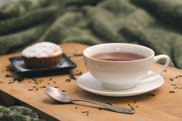 Homemade cupcake with powdered sugar on a black plate and a white cup of tea with natural additives on a wooden tray on a background of green textiles. The concept of a cozy breakfast in bed