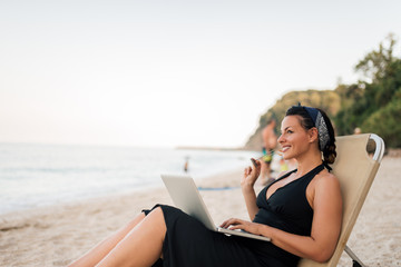 Portrait of a beautiful smiling young woman sitting on the beach with laptop.