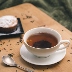 Homemade cupcake with powdered sugar on a black plate and a white cup of tea with natural additives on a wooden tray on a background of green textiles. The concept of a cozy breakfast in bed