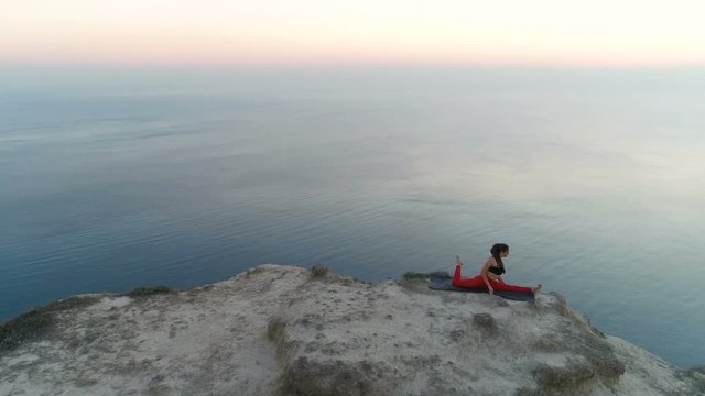Beautiful woman is doing yoga stretcing Pigeon Pose on the top of mountain with sea view at sunset. Aerial footage.