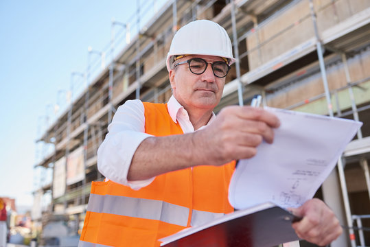 Mann, Architekt mit Helm auf Baustelle, mit Plan in der Hand und Haus im Hintergrund